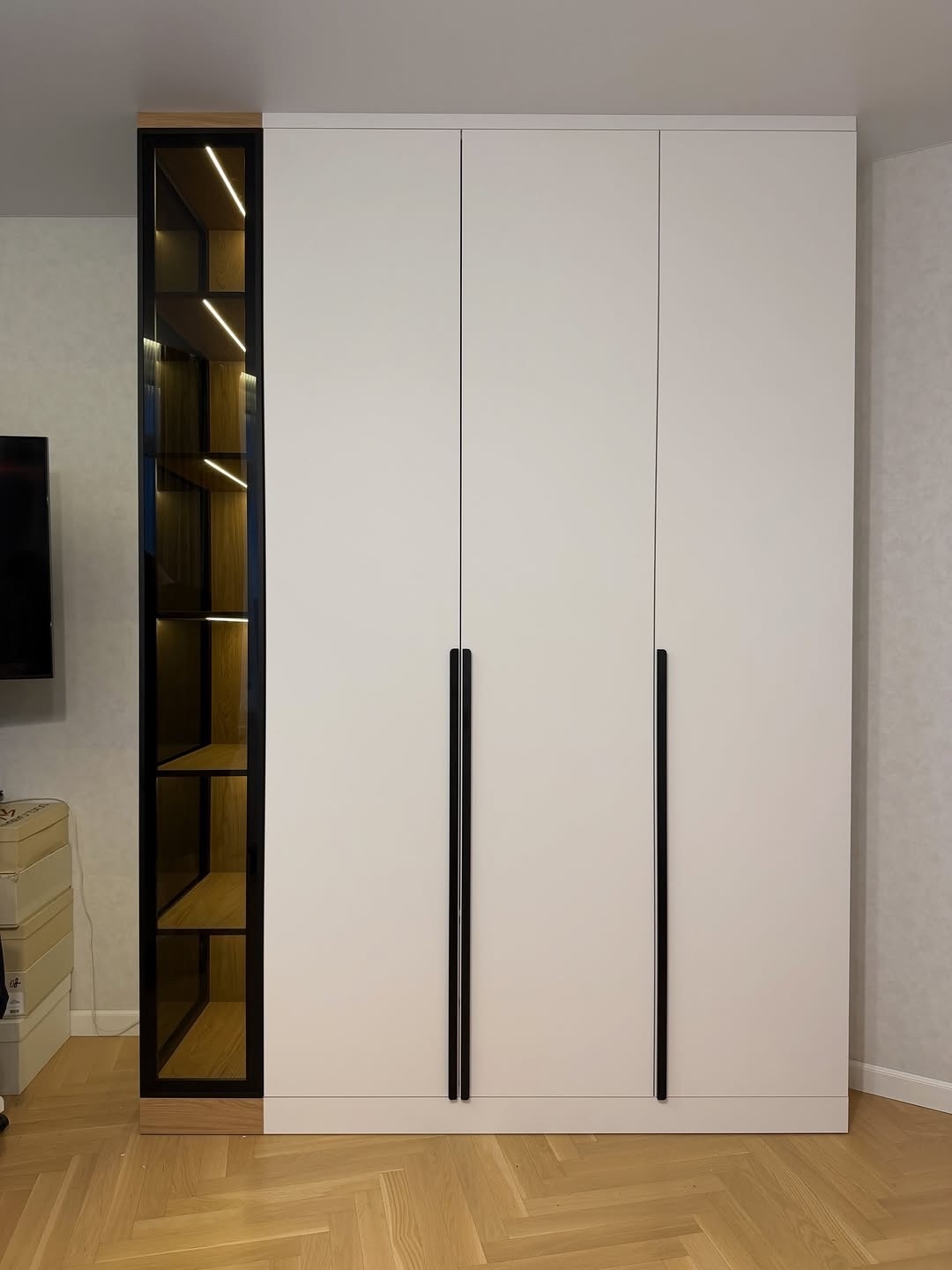Шкаф в закрытом виде: светлые фасады, длинные чёрные ручки и подсвеченная боковая витрина.